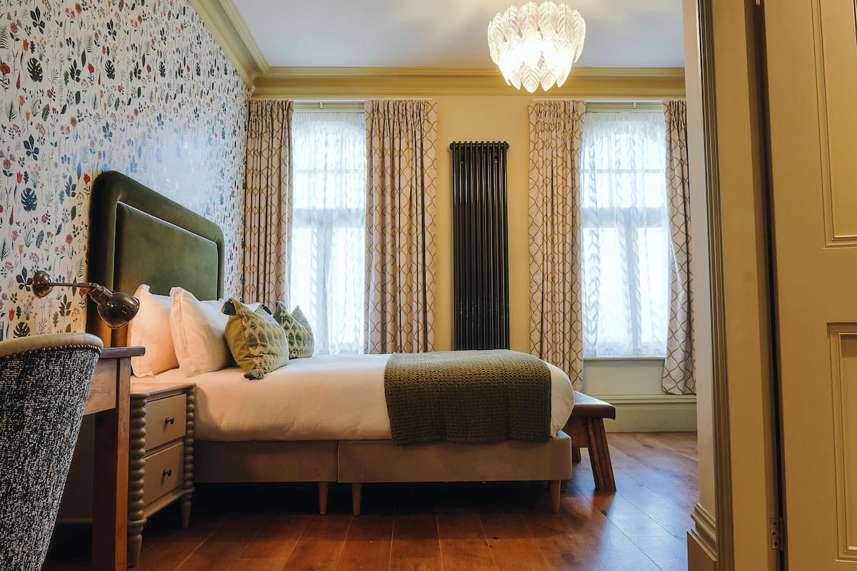 Quarto com cama, mesa de cabeceira, luminária e janela com cortinas.