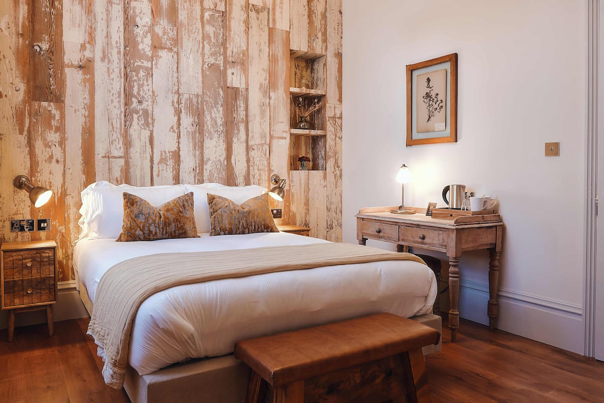 Quarto com parede de madeira, cama com almofadas, mesa de cabeceira com abajur, um banco de madeira e cômoda com espelho.