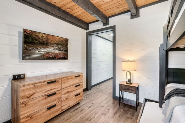 A bedroom with a wooden dresser, a bedside table with a lamp, a bed, and a large painting of a stream on the wall.