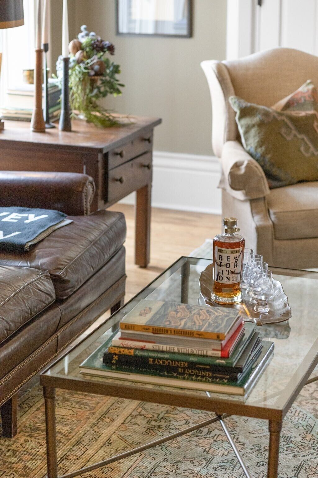 Un salón con un sofá de cuero, una mesa de centro de vidrio con libros y una botella de whisky, y un sillón beige.