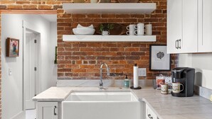 A kitchen with a brick wall, white cabinets, a marble countertop, and a coffee maker.