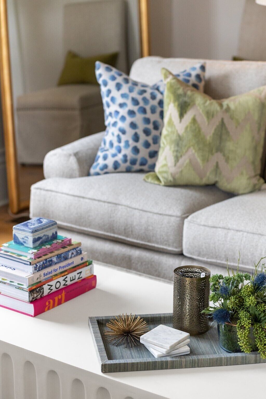 A living room with a grey sofa, patterned pillows, a stack of books, a tray with decorative items, and a small plant.