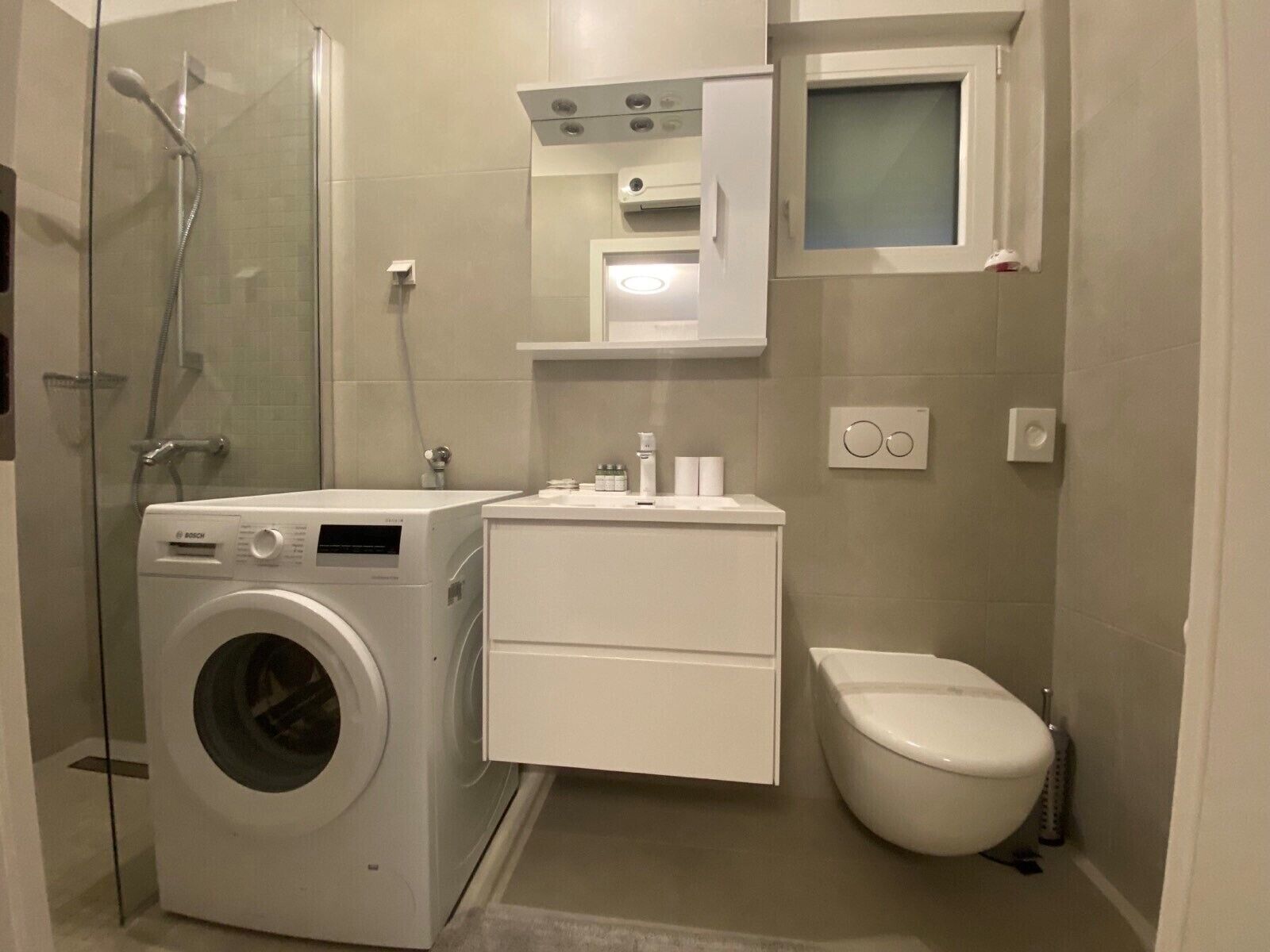 Ein kompaktes, modernes Badezimmer mit einer Bosch Waschmaschine, Toilette, Waschbecken und einer Duschkabine.