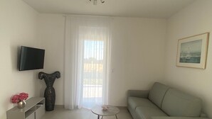 Un salon avec un canapé gris, une petite table ronde, un téléviseur fixé au mur et une fenêtre avec des rideaux blancs.