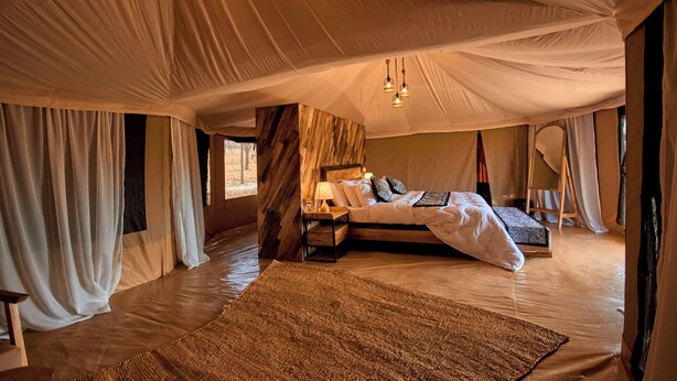 A tented room with a bed, a nightstand, a lamp, and a large woven rug. - Moyo Tented Camp (Serengeti)