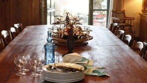 A wooden dining table with a central floral arrangement, surrounded by chairs, and a window with curtains in the background.