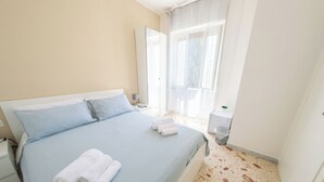 A neatly made bed with blue bedding and white pillows, a bedside table with a lamp, and a window with sheer curtains. - A Casa Di Marco (Salerno)