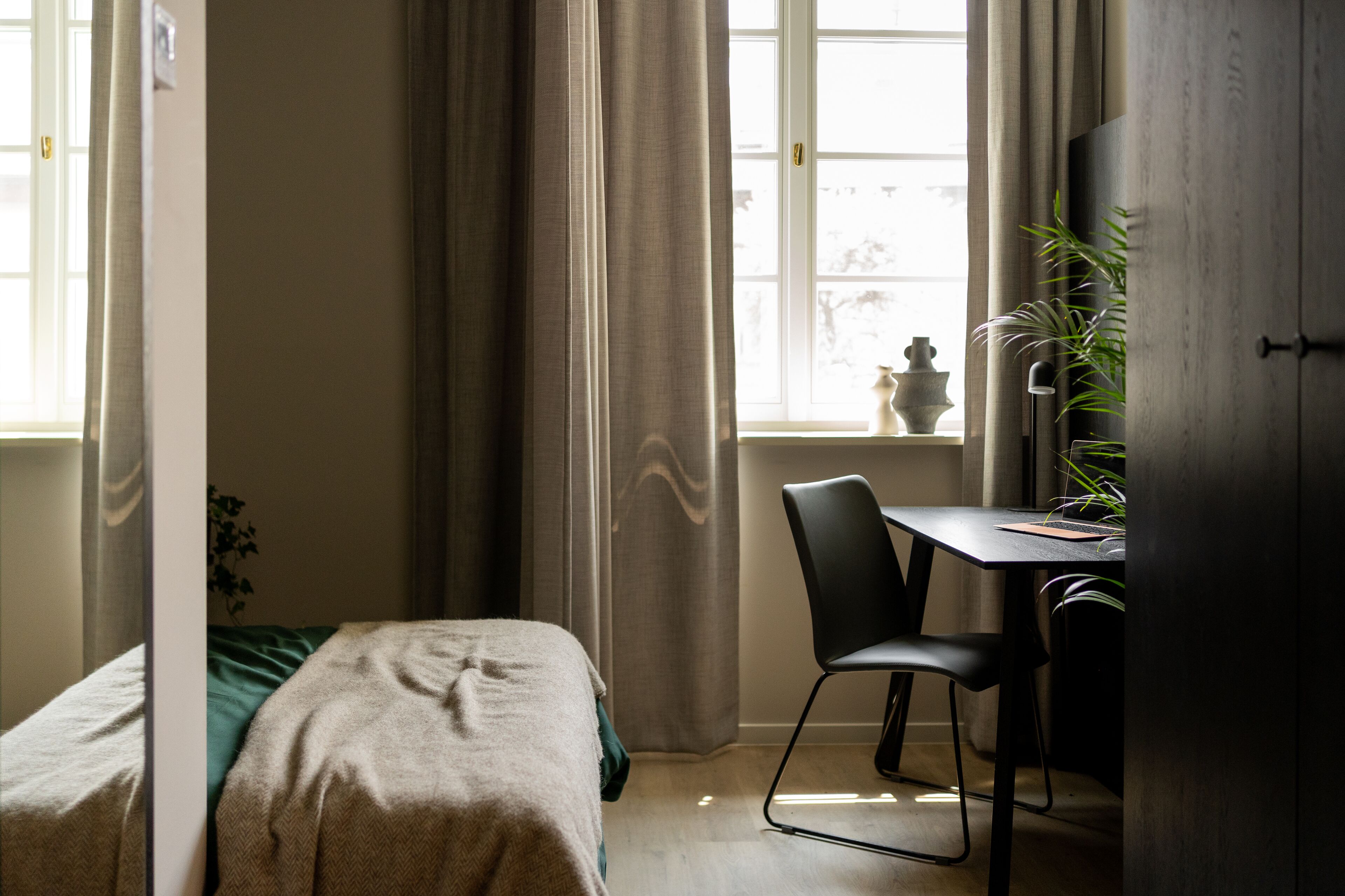 Ein Schlafzimmer mit einem Bett, einem Schreibtisch und einem Stuhl. Es gibt ein Fenster mit Vorhängen, eine Pflanze und einen Schrank.