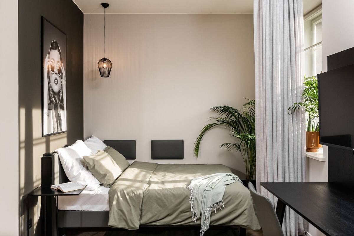 Ein modernes Schlafzimmer mit Bett, Nachttischen, einer Hängeleuchte, einem großen Fenster mit Vorhängen und einer Topfpflanze.