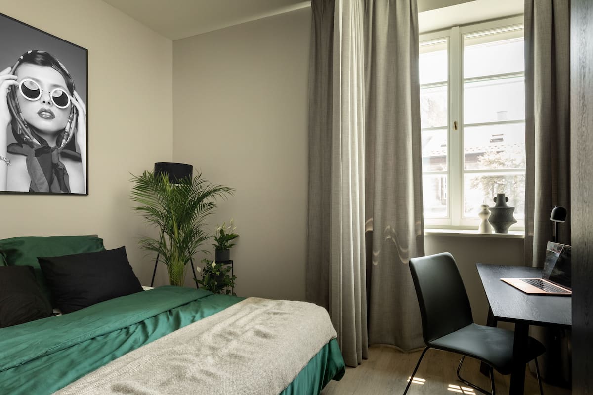 Ein Schlafzimmer mit einem Bett, einem Schreibtisch, einem Stuhl, einem Fenster mit Vorhängen und einer Topfpflanze.