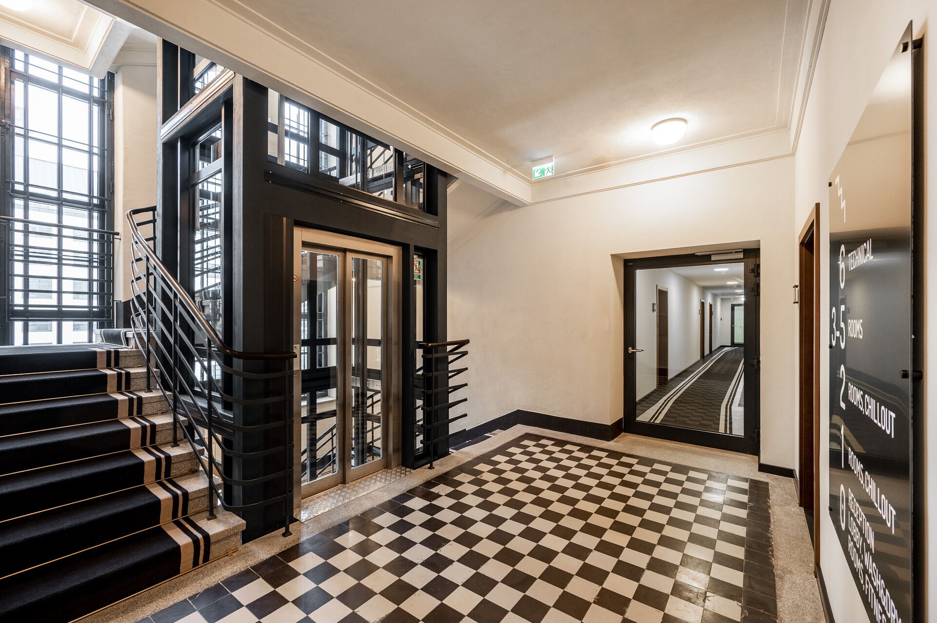 A hotel lobby with a checkered floor, a staircase with black railings, and a corridor leading to rooms.