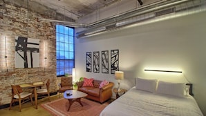 Una habitación de hotel moderna con paredes de ladrillo visto, un ventanal grande, una cama, un sofá, una mesa de centro y obras de arte en las paredes.
