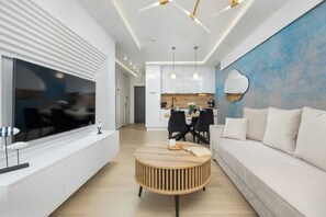 Una sala de estar moderna con televisor, mesa redonda de madera, sofá blanco y una cocina al fondo.