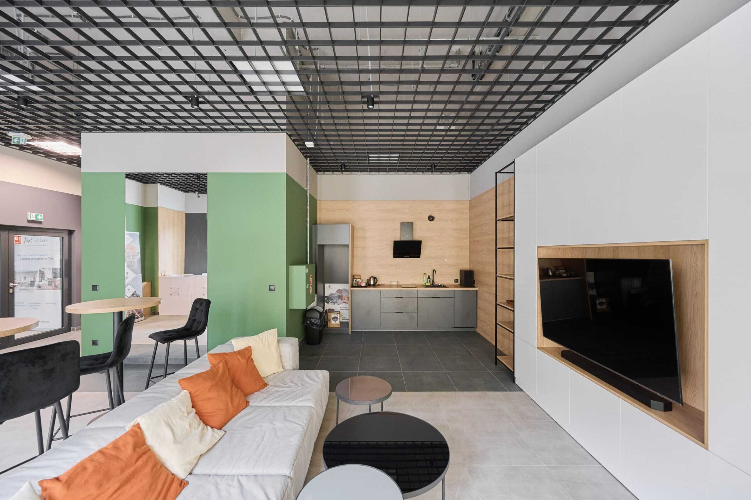 A modern hotel room with a sofa, a small round table, a TV mounted on the wall, and a kitchenette area.