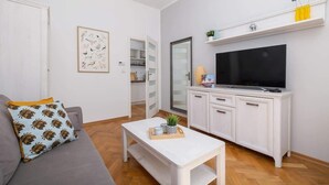 Una sala de estar moderna con un sofá gris, una mesa de centro blanca, un televisor de pantalla plana y una cocina visible a través de una puerta abierta.