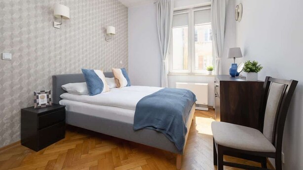 Un dormitorio con cama, mesita de noche, silla y ventana con cortinas.