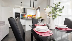 Un espace repas moderne avec une table en verre pour deux personnes, des chaises noires et un vase rempli de fleurs blanches. On aperçoit, à l’arrière-plan, une cuisine avec des meubles blancs et un mur décoratif aux couleurs vives.