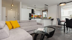 Un salon moderne avec un canapé gris, des coussins jaunes, une table basse en verre et un coin repas comprenant des chaises noires et une table en verre.