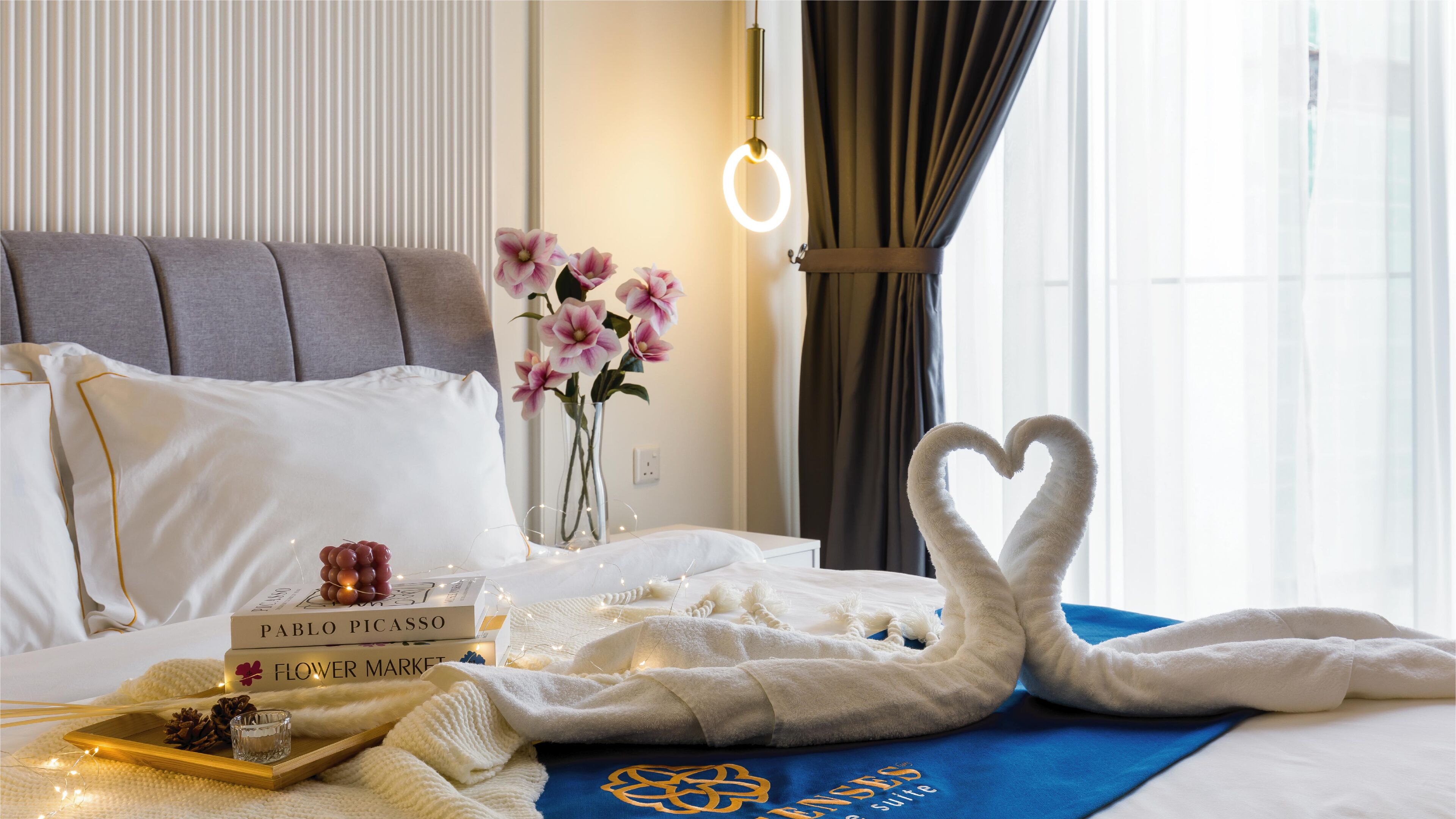 Een net opgemaakt bed met witte lakens, een handdoek opstelling in de vorm van een hart, een nachtkastje met een boek getiteld 'Pablo Picasso' en 'Bloemenmarkt', en een vaas met roze bloemen.