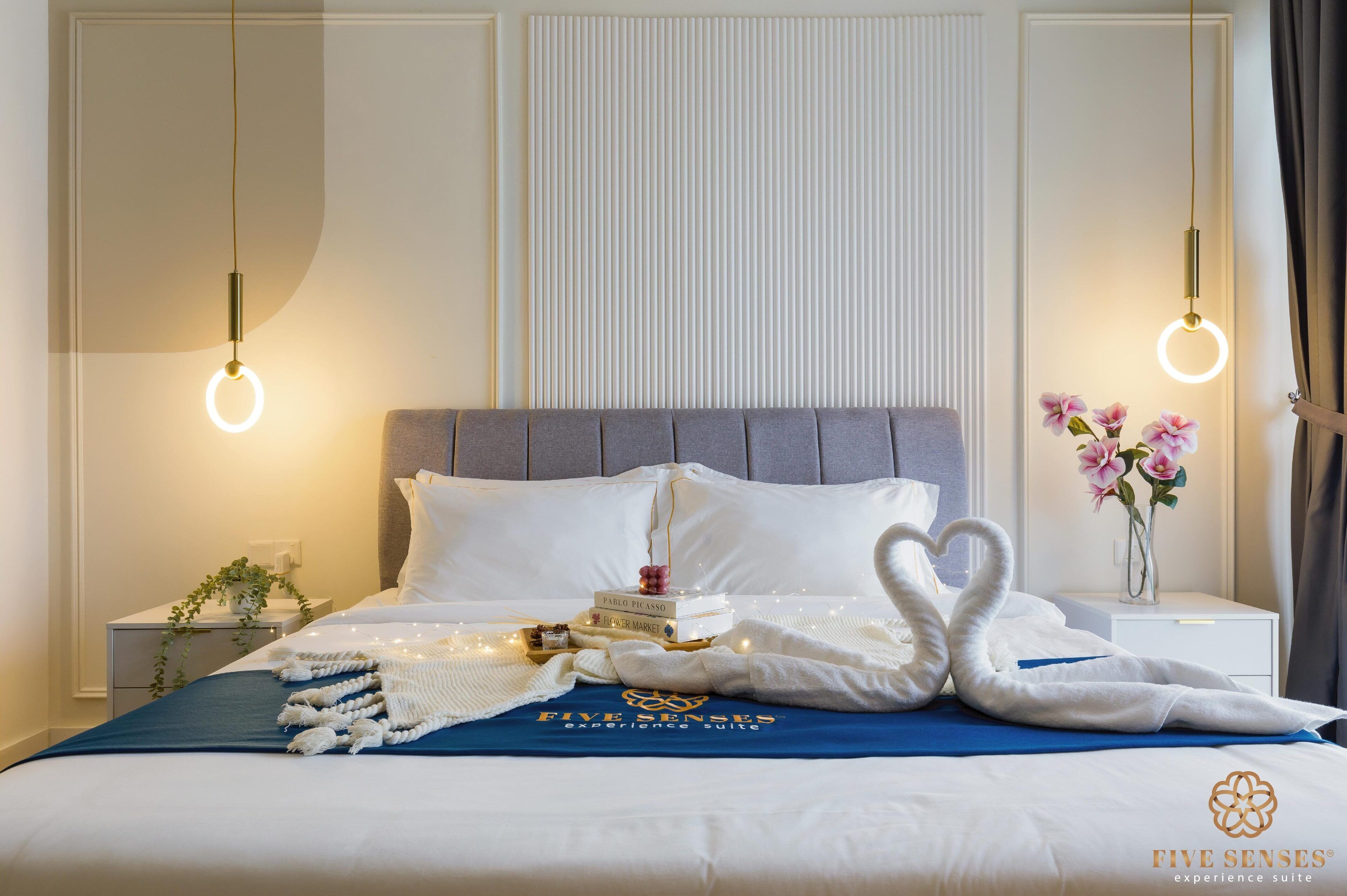 Un lit bien fait, recouvert d’une couverture bleue, agrémenté d’une pile de serviettes pliées en forme de cœur, d’une pile de livres et d’un vase rempli de fleurs roses.