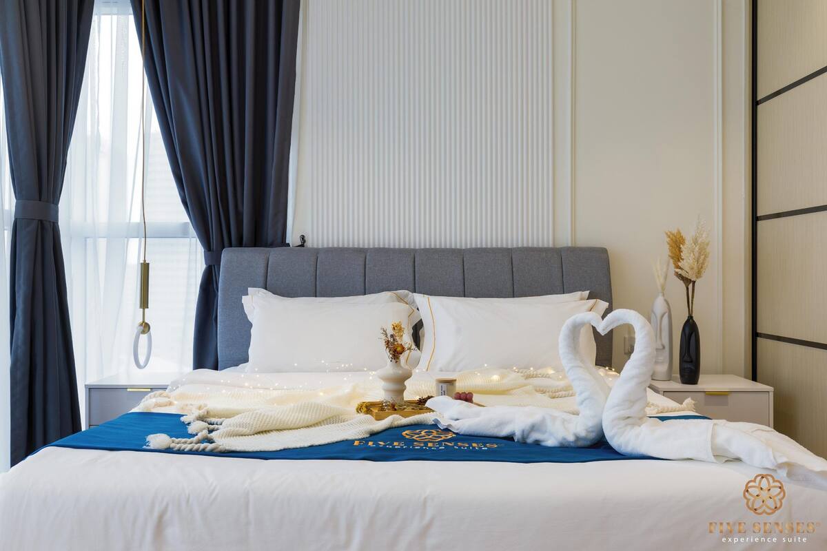 Een net opgemaakt bed met witte lakens, een blauwe deken en decoratieve zwanen van handdoeken. Op het nachtkastje naast het bed staat een vaas met gedroogde bloemen.