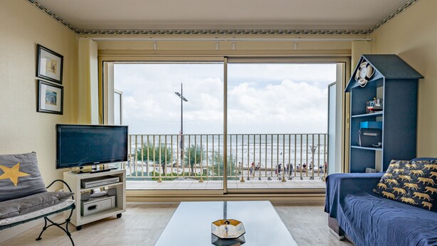 Ein Wohnzimmer mit Fernseher, Sofa und Couchtisch, ein Balkon mit Blick auf den Strand und Personen.