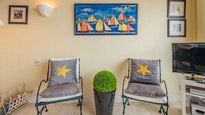 Ein Wohnzimmer mit zwei Sesseln, einem runden Tisch mit einer grünen Kugelgestalt, einem Segelbootgemälde und einem Fernseher auf einem Schrank.