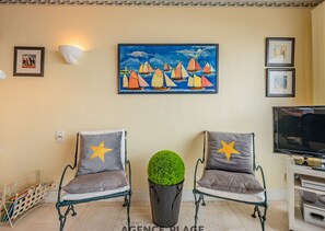 Una sala de estar con dos sillones, una mesa redonda con un topiario verde, un cuadro de veleros y un televisor sobre un mueble.