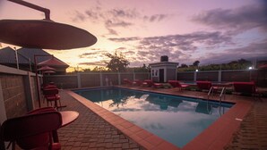 Una piscina con sillas reclinables rojas y un pequeño edificio, rodeado por un muro de ladrillo y un área de patio empedrada.