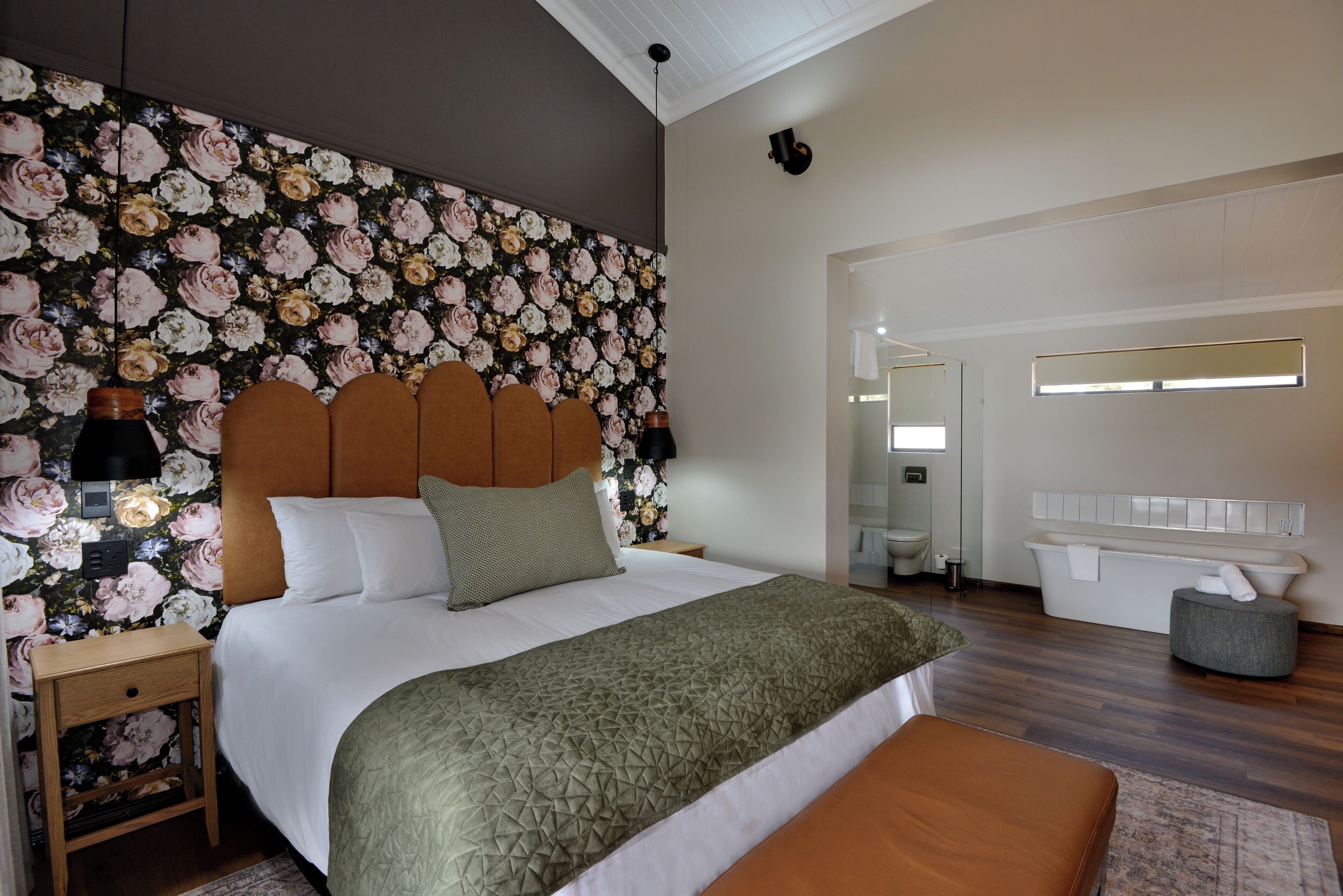Een slaapkamer met bloemenbehang, een houten hoofdbord, een bed met witte lakens, een groene plaid, een nachtkastje met een lamp, en een badkamer met een witte badkuip en een spiegel.