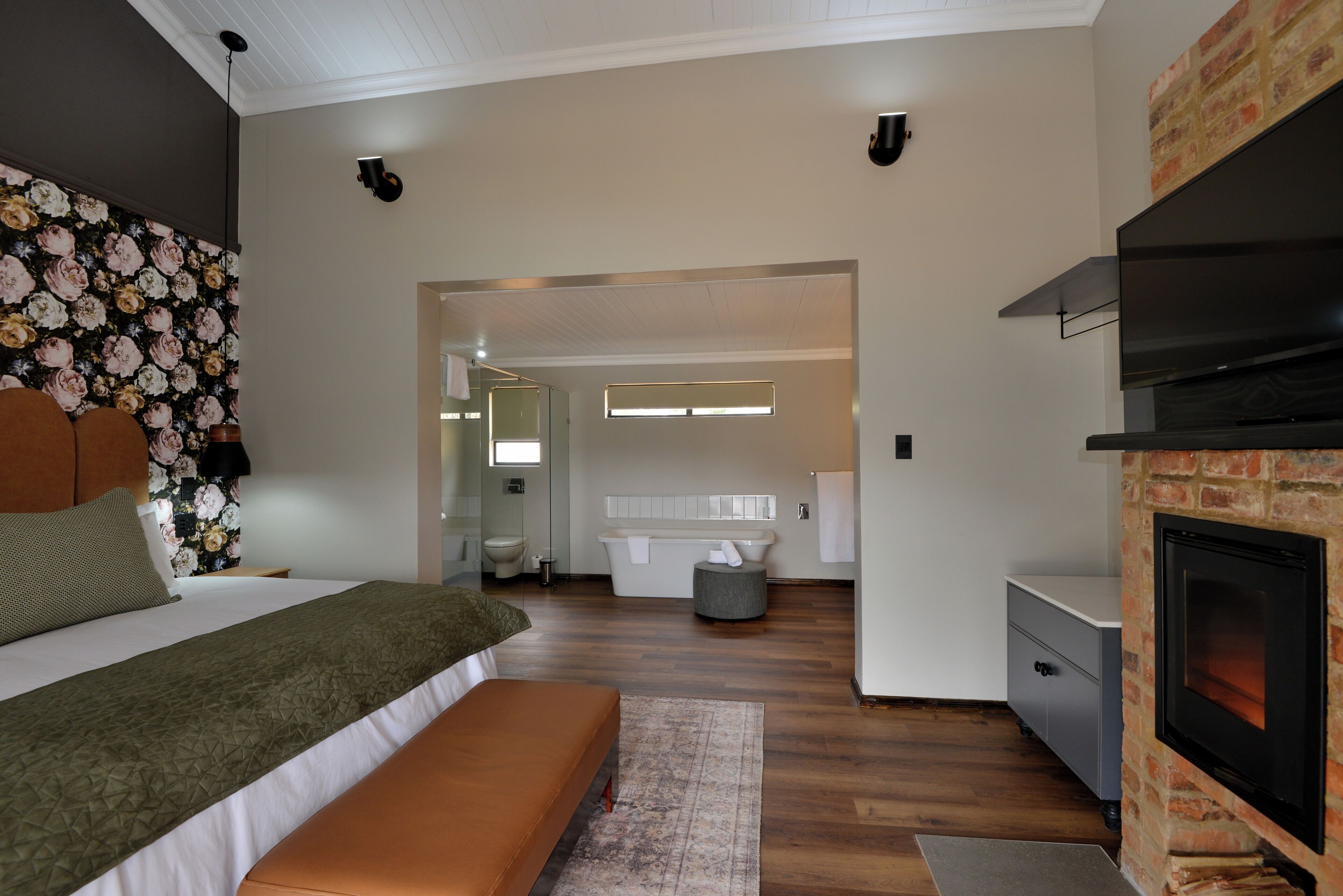Een moderne slaapkamer met een groot bed, een bankje, een open haard en een badkamer op de achtergrond.