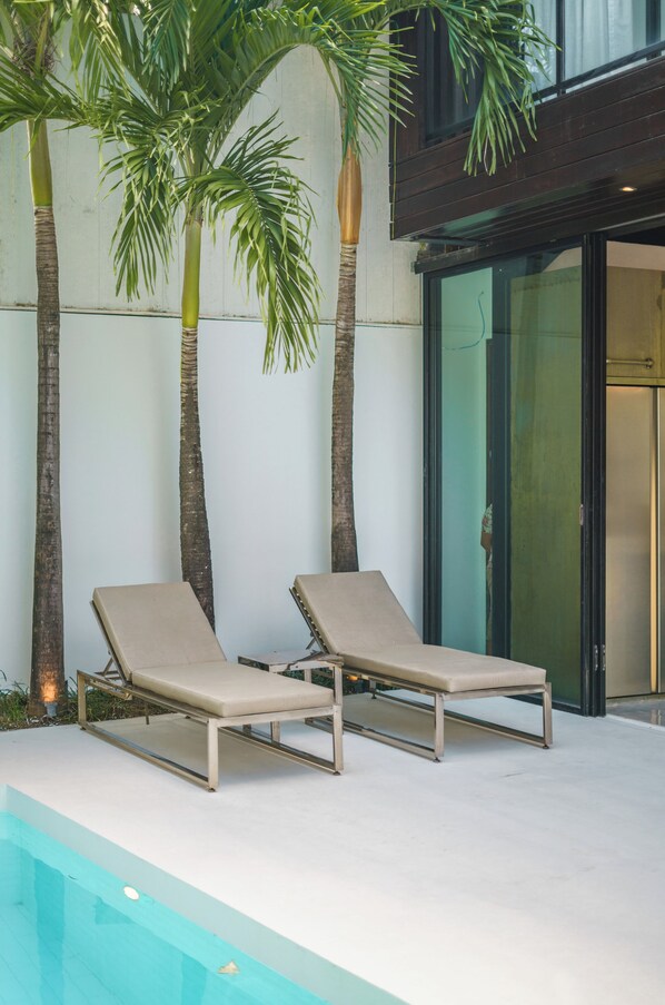 Un espace aménagé au bord de la piscine, avec deux chaises longues, des palmiers et un bâtiment moderne doté de portes vitrées.