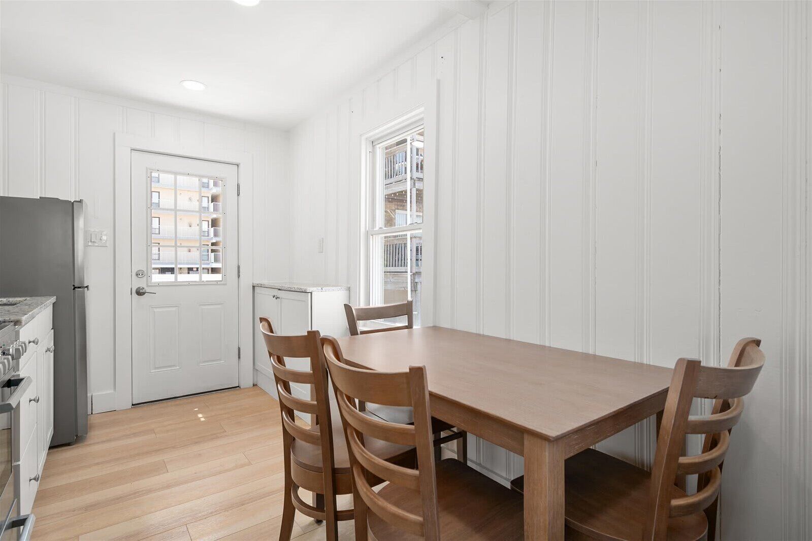 A dining area with a wooden table and chairs, a white door, and a view of balconies through a window.