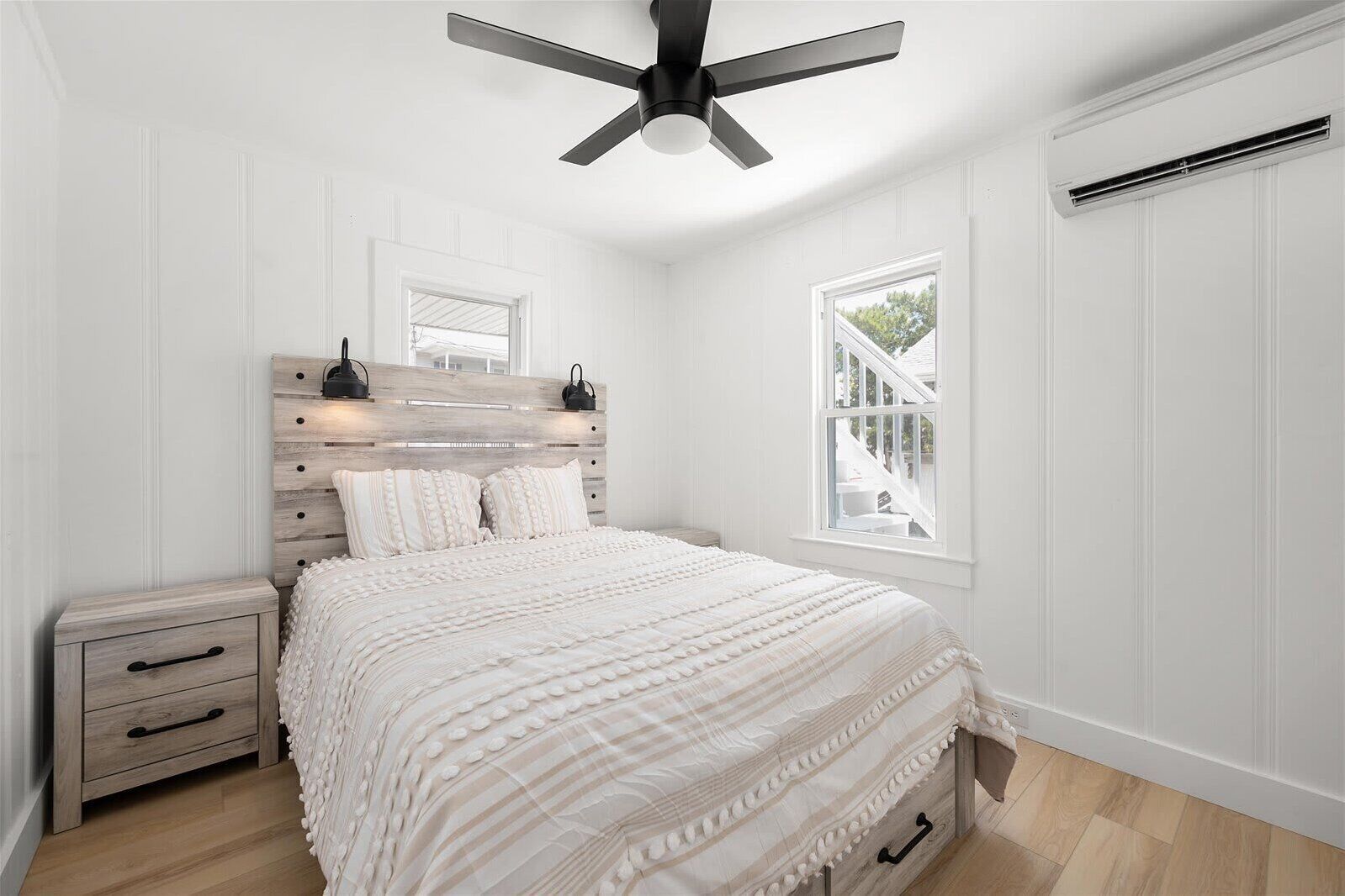 A bedroom with a wooden headboard, a bed with white bedding, a nightstand, a drawer, a ceiling fan, and a window with a view of outdoor stairs.