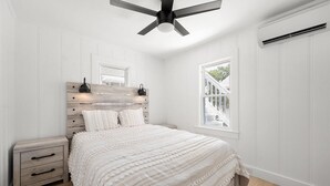 A bedroom with a wooden headboard, a bed with white bedding, a nightstand, a drawer, a ceiling fan, and a window with a view of outdoor stairs.