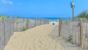 Un camino de arena con valla de madera que conduce a una zona de playa con sombrillas y gente.
