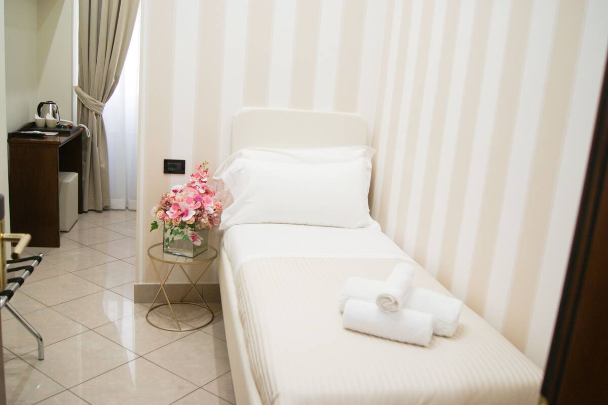 Ein Hotelzimmer mit einem Einzelbett, einem Nachttisch mit Blumenvase und einem Sessel.