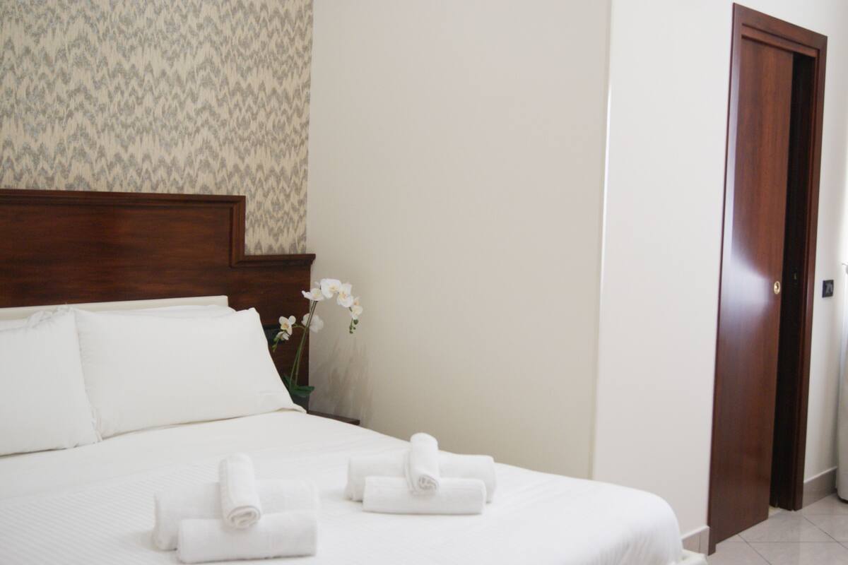 Ein Hotelzimmer mit einem ordentlich bezogenen Bett, einem hölzernen Kopfteil, einer Vase mit Blumen und einer geschlossenen Tür.