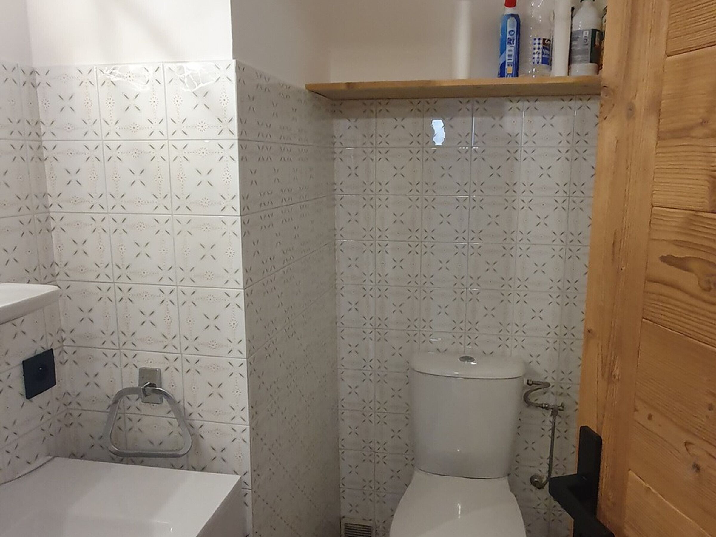 Kylpyhuoneessa on kuvioituja laattoja, wc-istuin, kaide ja hylly, jossa on hygieniatuotteita.