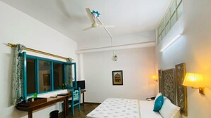 A hotel room with a bed, a desk, a chair, a ceiling fan, a window with curtains, a wall-mounted lamp, and a small table with a bottle. - Mango Tree Courtyard Dehradun (Dehradun)