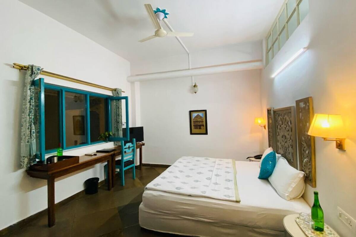 A hotel room with a bed, a desk, a chair, a ceiling fan, a window with curtains, a wall-mounted lamp, and a small table with a bottle.