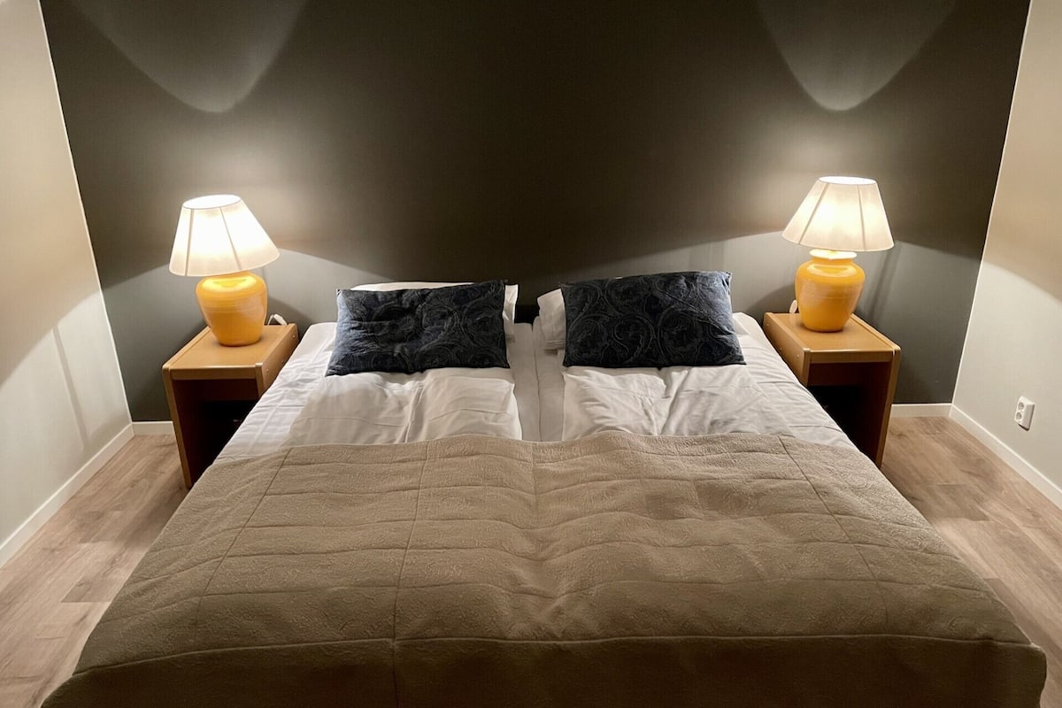 A bed with a beige quilt, two dark pillows, and two bedside lamps on wooden nightstands.