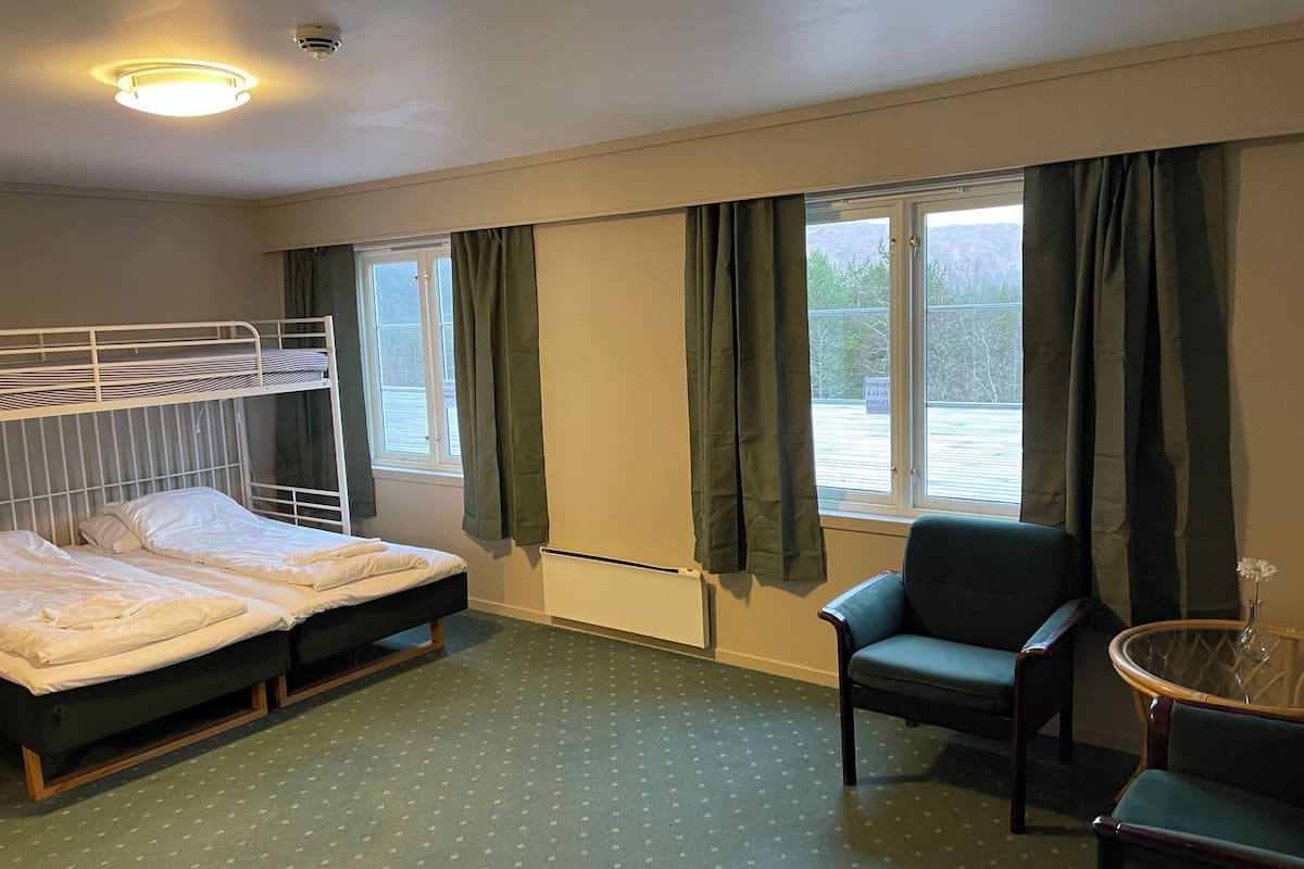 A hotel room with a bunk bed, a single bed, a chair, a small table, and a window with curtains.