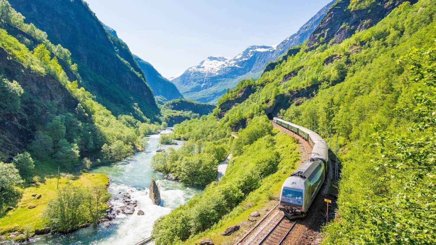 Un tren travessa un paisatge exuberant amb un riu i muntanyes al fons.