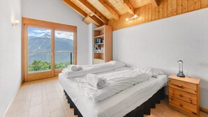 Une chambre avec une grande fenêtre, des poutres apparentes en bois au plafond, un lit, une table de chevet et une bibliothèque.
