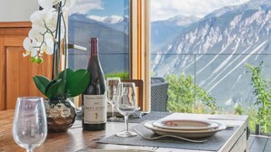 Une table à manger dressée avec des assiettes, des verres à vin et une bouteille de vin, avec une vue sur les montagnes par une grande fenêtre.