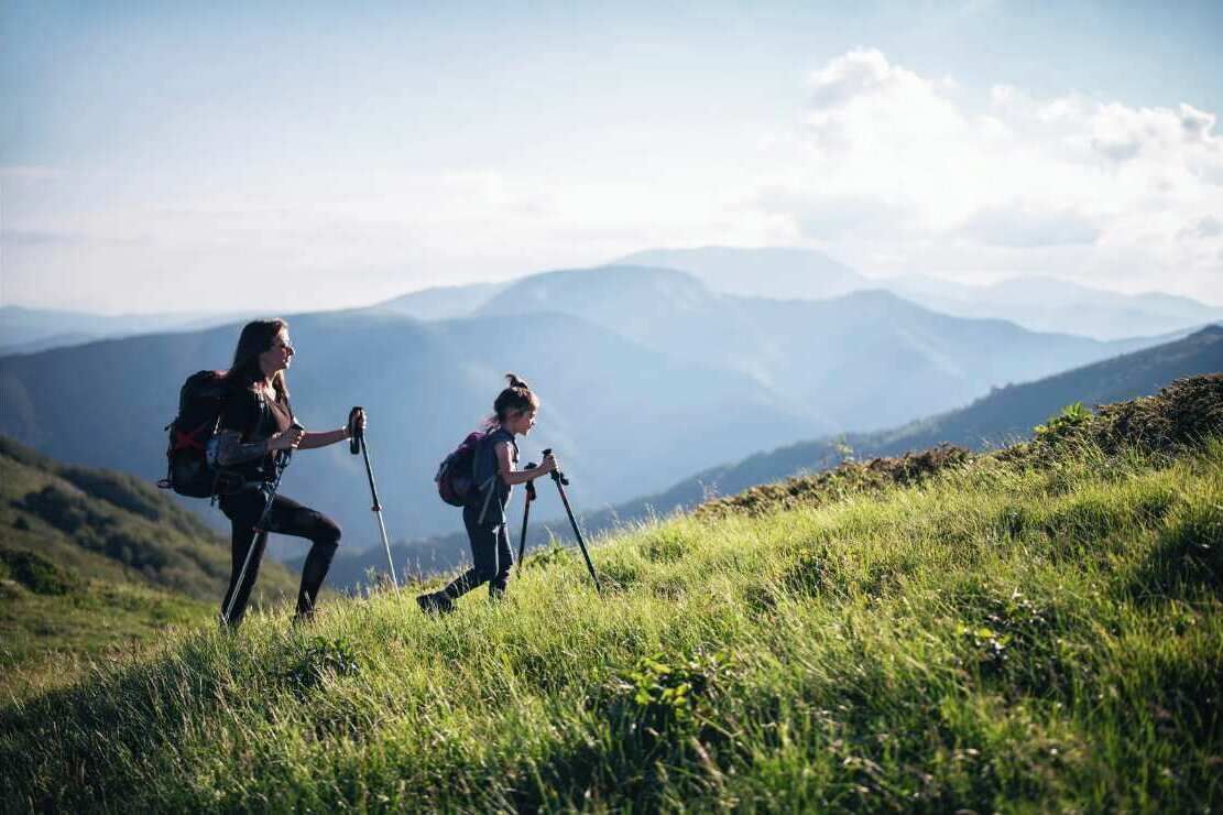 Dos excursionistas en un sendero montañoso cubierto de hierba, con montañas distantes al fondo.