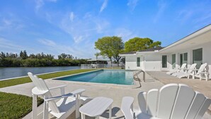 A pool area with white Adirondack chairs and tables, a clear view of a lake, and a building with large windows. - Luxury Beachside 4BR/3BA Waterfront Home (Cocoa Beach)