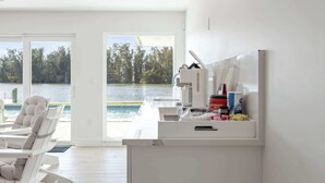 A modern kitchen with a large window overlooking a lake, a white chair, and a countertop with kitchen items. - Luxury Beachside 4BR/3BA Waterfront Home (Cocoa Beach)
