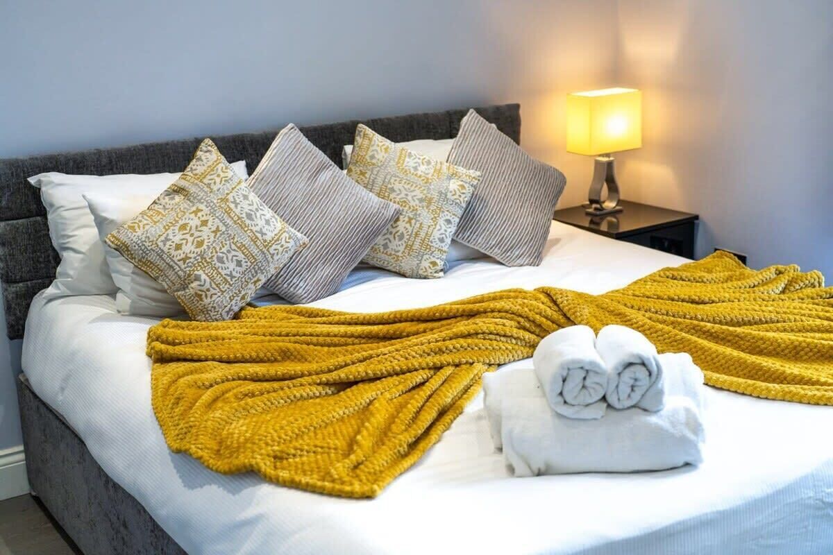 Uma cama bem arrumada com um cobertor amarelo, almofadas estampadas e toalhas dobradas.